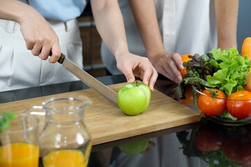 Closeup hand holding knife cutting green apple on a wooden chop board. Jar with mixed fruit juice and glass bowl with a variety of fruits and vegetables is placed on the kitchen counter.