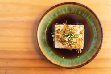 A dish of chilled tofu garnished with fried onions, sesame seeds, and green onions, served in soy sauce.