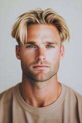 Fototapeta premium Young man with stylish blonde hair and blue eyes poses confidently against a neutral backdrop in a casual outfit during a personal photo session