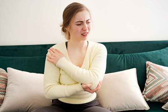 A young white woman holding injured elbow sitting on sofa indoor in the living room at home. Girl feeling pain in cubit area indoor. Discomfort, joint injury, chronic tendon arthritis, health problem.