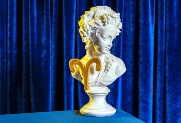 A female plaster bust with the golden sign of Aries on the background of blue velvet curtains still life.