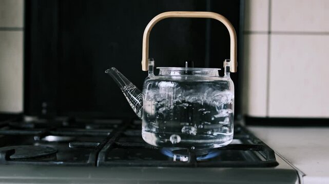 Glass kettle with a wooden handle boiling water on a gas stove, with visible bubbles indicating the heat and steam. The transparent glass showcases the boiling process and kitchen setting.