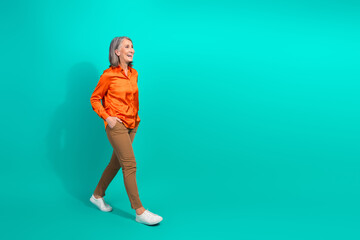 Mature woman walks confidently wearing a vibrant orange shirt and brown pants on a teal backdrop for lifestyle promotion purposes