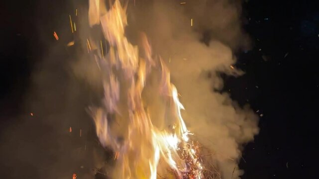 Bright firecamp with sparks at night. Fire pit outdoors, wood and dry grass on fire, flying sparks and smoke.