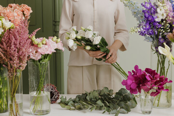 Female florist creating a delicate bouquet with fresh flowers in a floral workshop, surrounded by colorful blooms in glass vases. flower shop