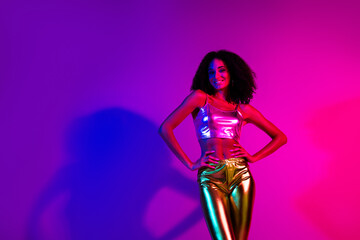 Fashionable young woman posing in vibrant lighting featuring neon pink, purple, and blue tones, showcasing stylish metallic attire