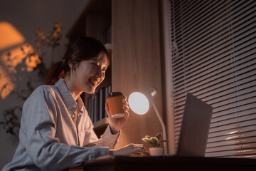 Asian businesswoman working from home at night in her cozy home office, sipping coffee while using her laptop, illuminated by a warm desk lamp, creating a comfortable atmosphere