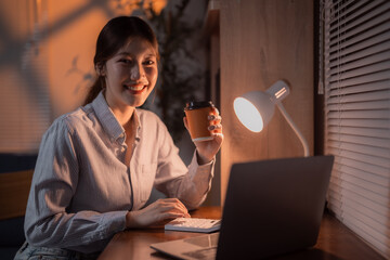 Young Asian businesswoman drinking coffee and working late at night in her home office, using calculator and laptop under a desk lamp light