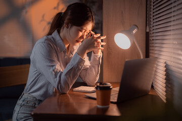 Young office worker massaging her temples because of a strong headache, sitting at her desk with laptop and calculator, working late in the evening