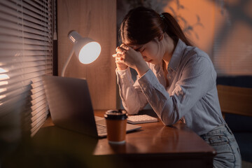 Young Asian office worker feeling tired and stressed while working late at night on laptop...