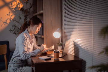 Asian businesswoman writing on notebook while using laptop computer working from home at night,...