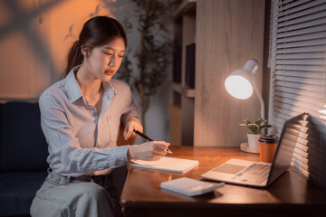 Concentrated Asian businesswoman writing notes while using laptop computer, working late at night...