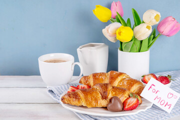 Mother’s Day breakfast background featuring croissant, strawberries, chocolates, coffee mug, bouquet of tulip flowers, as surprise and gift, with greeting card 