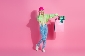 Happy young woman shopping with colorful bags, wearing trendy casual outfit and beanie hat, standing against pink background