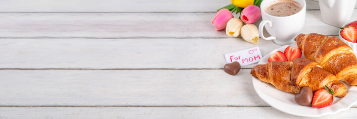 Mother’s Day breakfast background featuring croissant, strawberries, chocolates, coffee mug, bouquet of tulip flowers, as surprise and gift, with greeting card 
