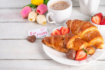 Mother’s Day breakfast background featuring croissant, strawberries, chocolates, coffee mug, bouquet of tulip flowers, as surprise and gift, with greeting card 