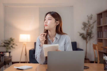 Young Asian businesswoman holding a pen and notebook, thinking deeply while working on a laptop in...