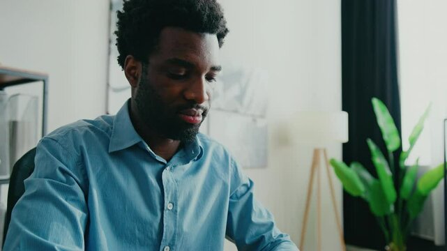 Cheerful African American man in blue shirt sitting at desk. Operating keyboard while looking at computer screen. Engaging in productive work or enjoying online communication in bright modern office.