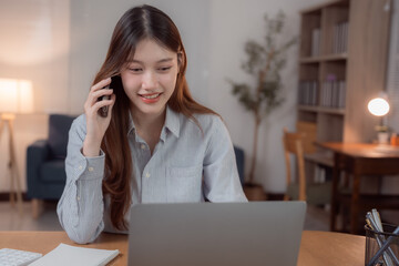 Smiling asian businesswoman working from home, using laptop computer and talking on mobile phone,...