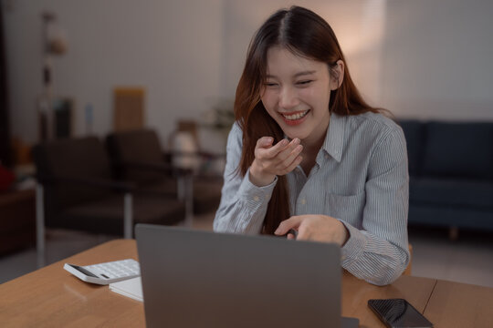Happy young Asian businesswoman is making a video conference on laptop, talking, laughing and gesturing with hand sitting at wooden table with calculator and smartphone in living room at home