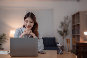 Young Asian business woman working from home using laptop computer smiling and looking at screen,...