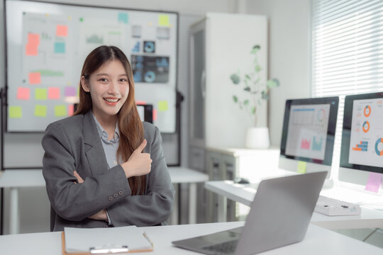 Young asian businesswoman showing thumbs up with crossed arms while sitting at desk and working with laptop and desktop computer in modern office, smiling and looking satisfied with her work