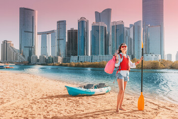 Young woman enjoys kayaking in front of Abu Dhabi skyline, holding a paddle and a waterproof bag