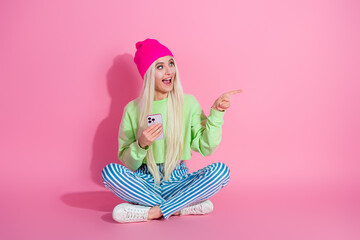 Young stylish woman in vibrant casual outfit sitting on pink background pointing cheerfully, holding smartphone