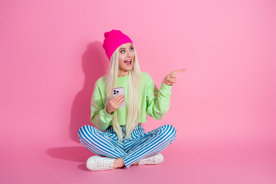 Young stylish woman in vibrant casual outfit sitting on pink background pointing cheerfully, holding smartphone