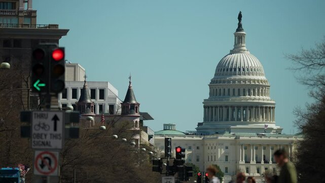 Red and green traffic lights are visible as pedestrians navigate, with the United States Capitol building in Washington, DC standing prominently against a clear sky.