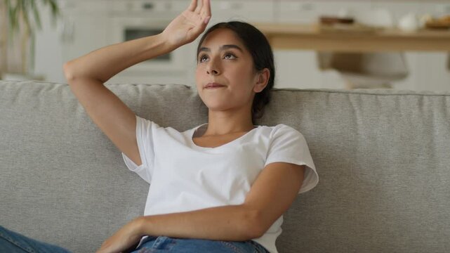 Sad upset Indian woman feel overheated waving hands Arabian girl female suffering annoyed with high temperature inside living room home conditioner broken stressed sweaty on sofa summer weather heat