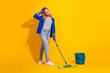 Young woman in casual stylish attire with mop and bucket on vibrant yellow themed background