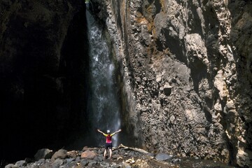 A small silhouette of a woman in a red T-shirt standing with her arms raised in front of a waterfall in Arusha National Park, Tanzania