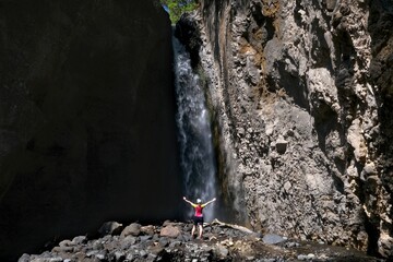 A small silhouette of a woman in a red T-shirt standing with her arms raised in front of a waterfall in Arusha National Park, Tanzania