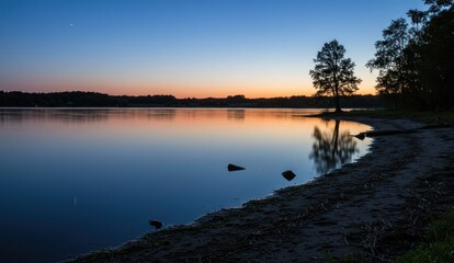 Obraz premium Calm lake reflects a colorful sunset sky, with trees silhouetted on the shore.