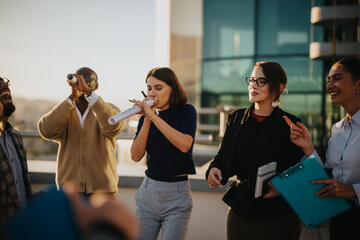 A diverse group of business people celebrates with energy on a rooftop balcony during sunset. The...