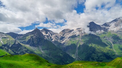 A beautiful landscape of snow-capped mountains rising above lush green hills. The sky is a deep blue with fluffy white clouds. A perfect backdrop for any outdoor adventure.