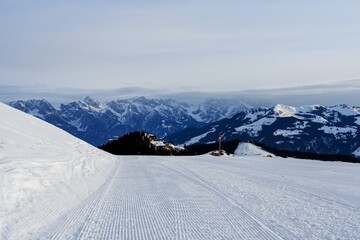 A stunning panoramic view of snow-covered mountain peaks under a clear blue sky. A groomed ski slope with fresh tracks invites skiers to enjoy this beautiful winter landscape.