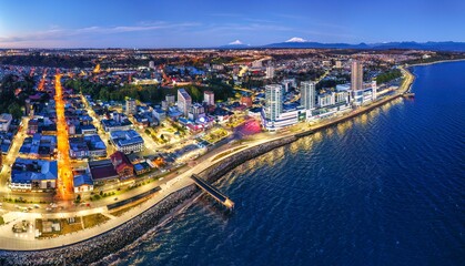 Naklejka premium Aerial drone view of Puerto Montt at twilight with a deep blue sky, faint orange sunset hues, and scattered clouds. The city glows with warm lights, and a serene waterfront, Osorno and Calbuco Volcano