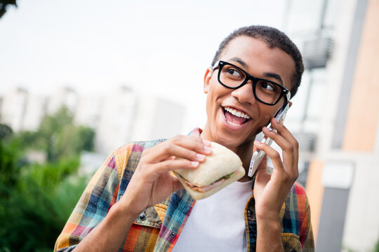 Photo of nice young man speak phone eat sandwich wear checkered shirt buildings city center outdoors
