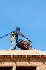 Workman in safety harness on wood framed house roof waiting for roofing materials being delivered by conveyor belt on a sunny winter day, home development construction project
