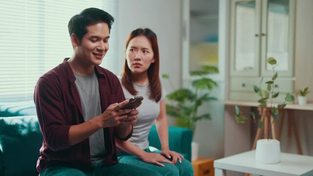 Young woman feeling jealous while her boyfriend is using his smartphone, sitting on the sofa at home