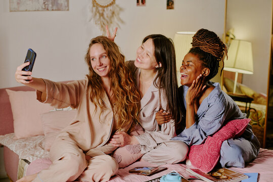 Three smiling biracial women in pajamas sitting together on bed and taking selfie to post on social media