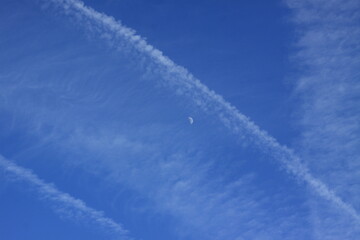 Blue sky featuring multiple intersecting airplane contrails, creating a crisscross pattern. The contrails are composed of white, wispy clouds that stretch horizontally across the image