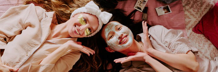 Website banner with two best friends with golden eye patches and clay mask on face lying on bed while posing for portrait