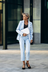 Fototapeta premium Elderly business woman holding laptop and standing in front of business building
