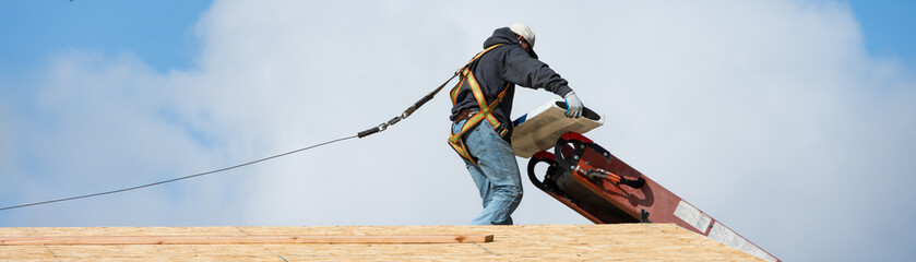Workman in safety harness on wood framed house roof carrying package of roofing materials delivered...