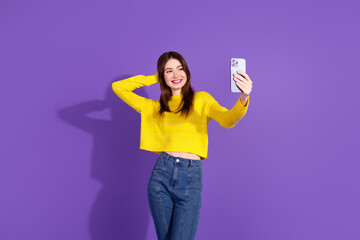 Young woman in a stylish yellow sweater enjoying a moment while taking a selfie against a vibrant purple backdrop