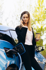 Business woman drinking coffee talking on the phone and standing by the electric car charging
