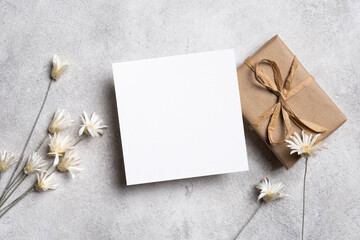 Blank square greeting card mockup with gift box and dried flowers, flat lay with copy space